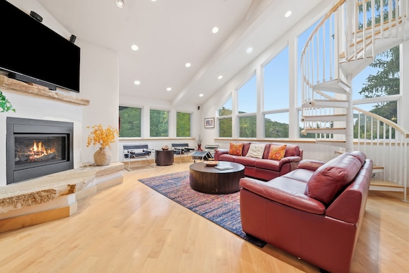 A spacious living room with a fireplace, leather sofas, a coffee table, and large windows offering a view of the outdoors.