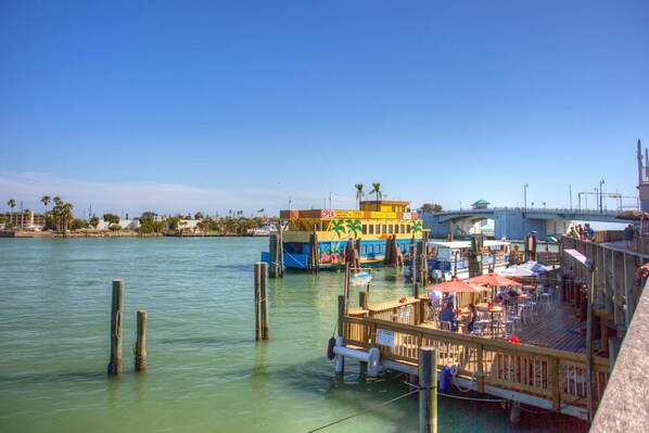 A waterfront restaurant with outdoor seating, a docked boat, and a bridge in the background.
