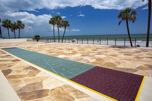 Una zona pavimentada con un camino táctil verde y púrpura, palmeras y una playa al fondo.