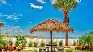 A thatched umbrella pavilion with a stone table and benches, surrounded by palm trees and flowering plants, under a clear blue sky.