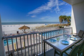 A balcony with a glass table and chairs overlooking a beach and a pool area.