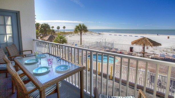 A balcony with a table set for dining, overlooking a beach with a pool and lounge chairs.