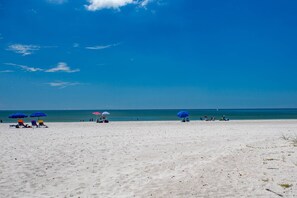 En strand med hvidt sand og en klar blå himmel. Flere parasoller og strandstole er placeret her.