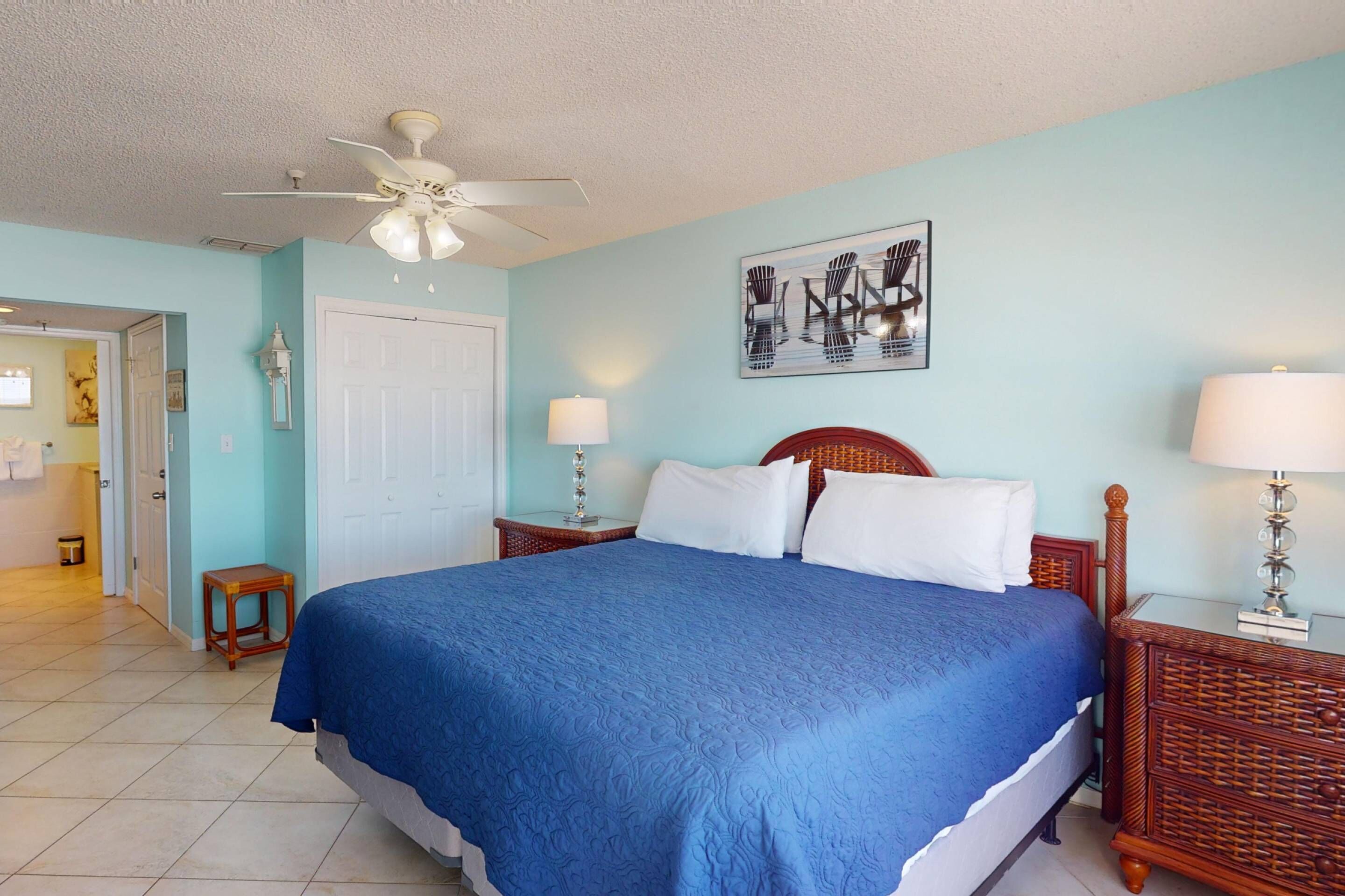 A bedroom with a large bed, two bedside tables, a ceiling fan, and a door leading to another room.