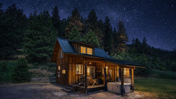 Una cabaña rústica con porche, zona de estar y jacuzzi, rodeada de un bosque y un cielo nocturno estrellado.