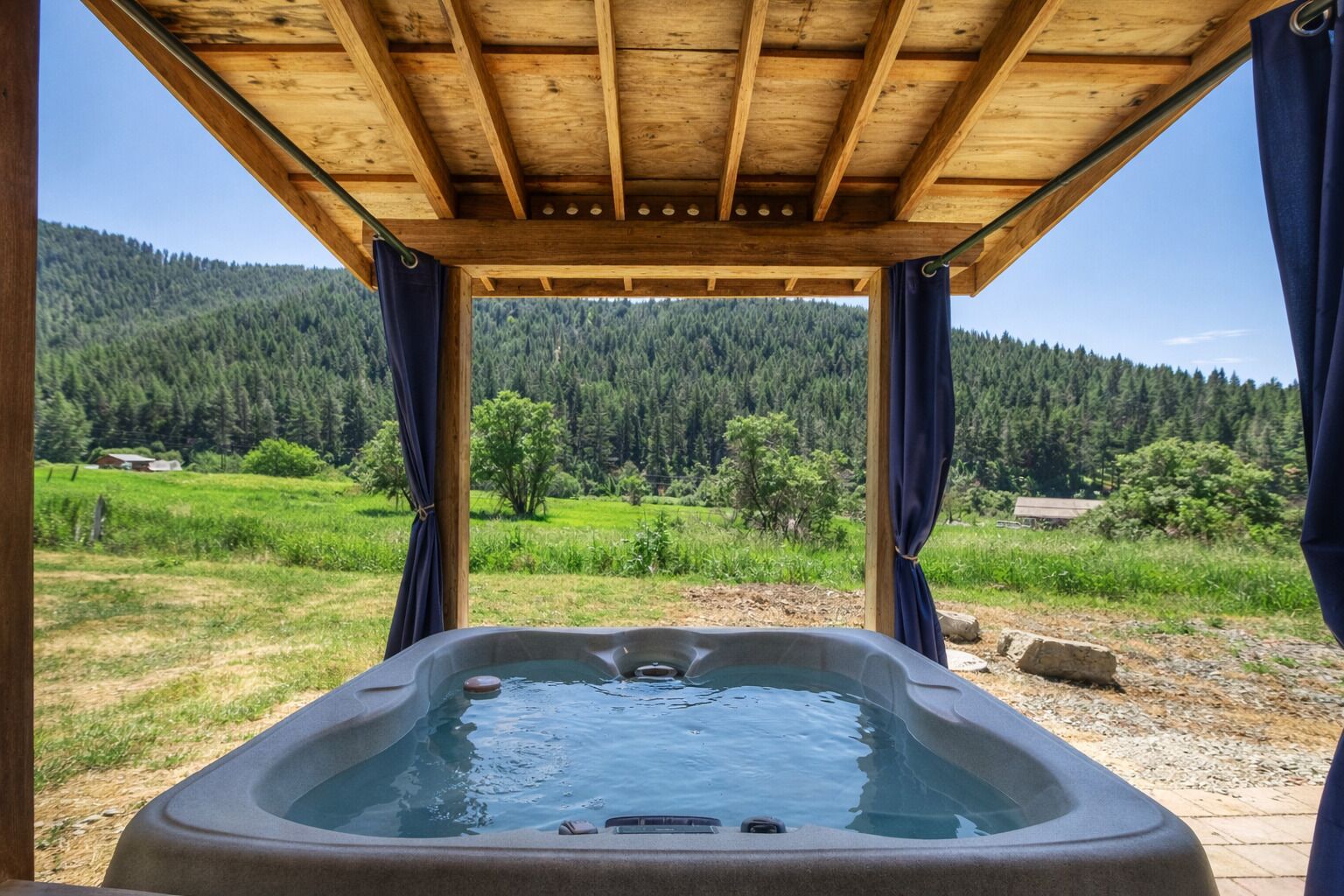 A hot tub under a wooden pergola with a view of a forested landscape.