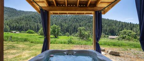 A hot tub under a wooden pergola with a view of a forested landscape.