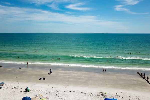 A beach with clear blue water, people swimming and walking on the sand, and a few umbrellas and beach chairs.