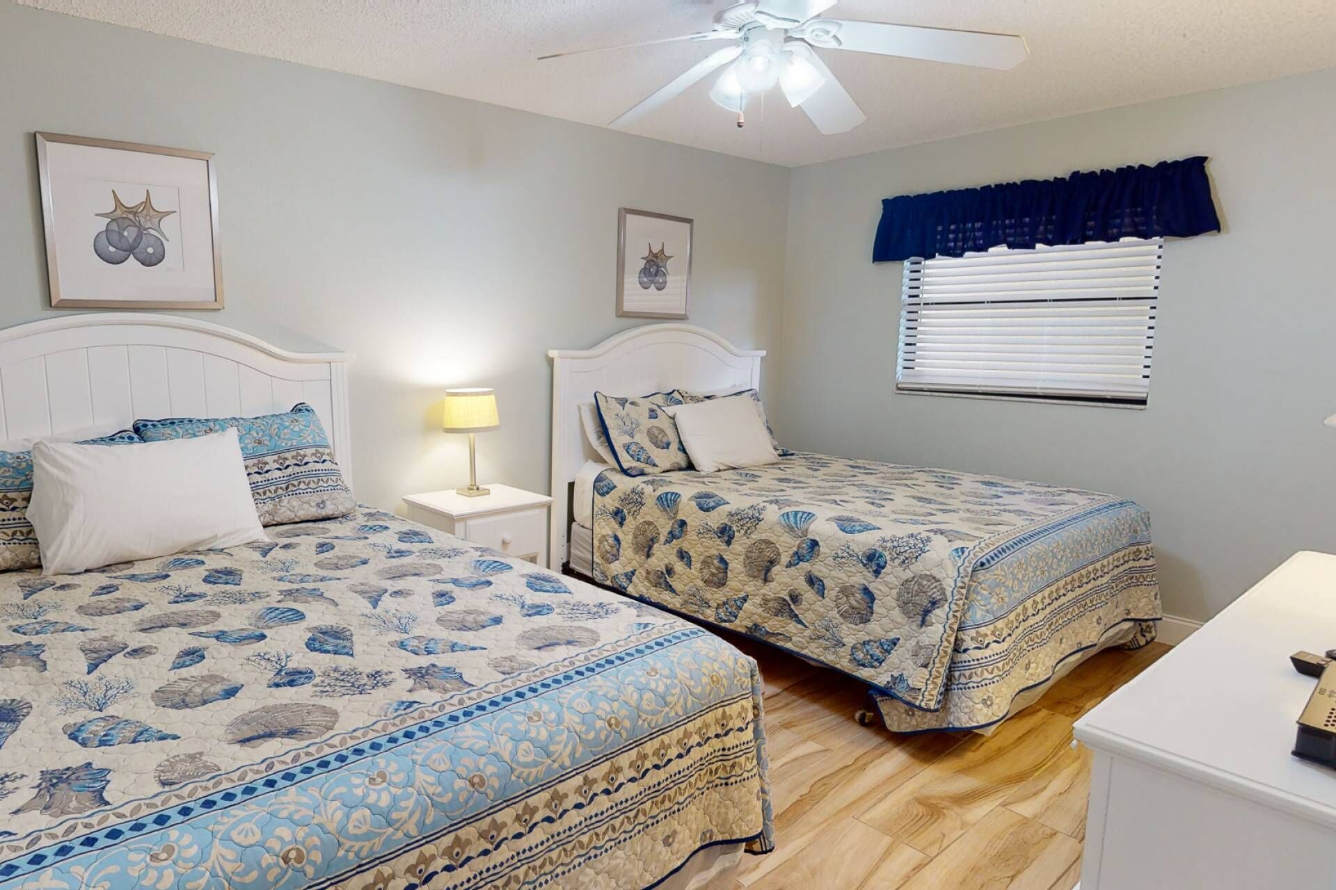 A bedroom with two beds, a ceiling fan, a window with blinds, and a nightstand with a lamp.