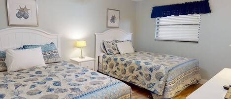 A bedroom with two beds, a ceiling fan, a window with blinds, and a nightstand with a lamp.