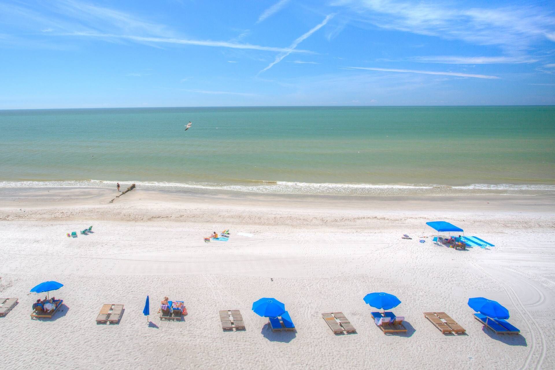 Ein Strand mit Liegestühlen, Sonnenschirmen, einer Mole, die ins Wasser ragt, und einem klaren Himmel.