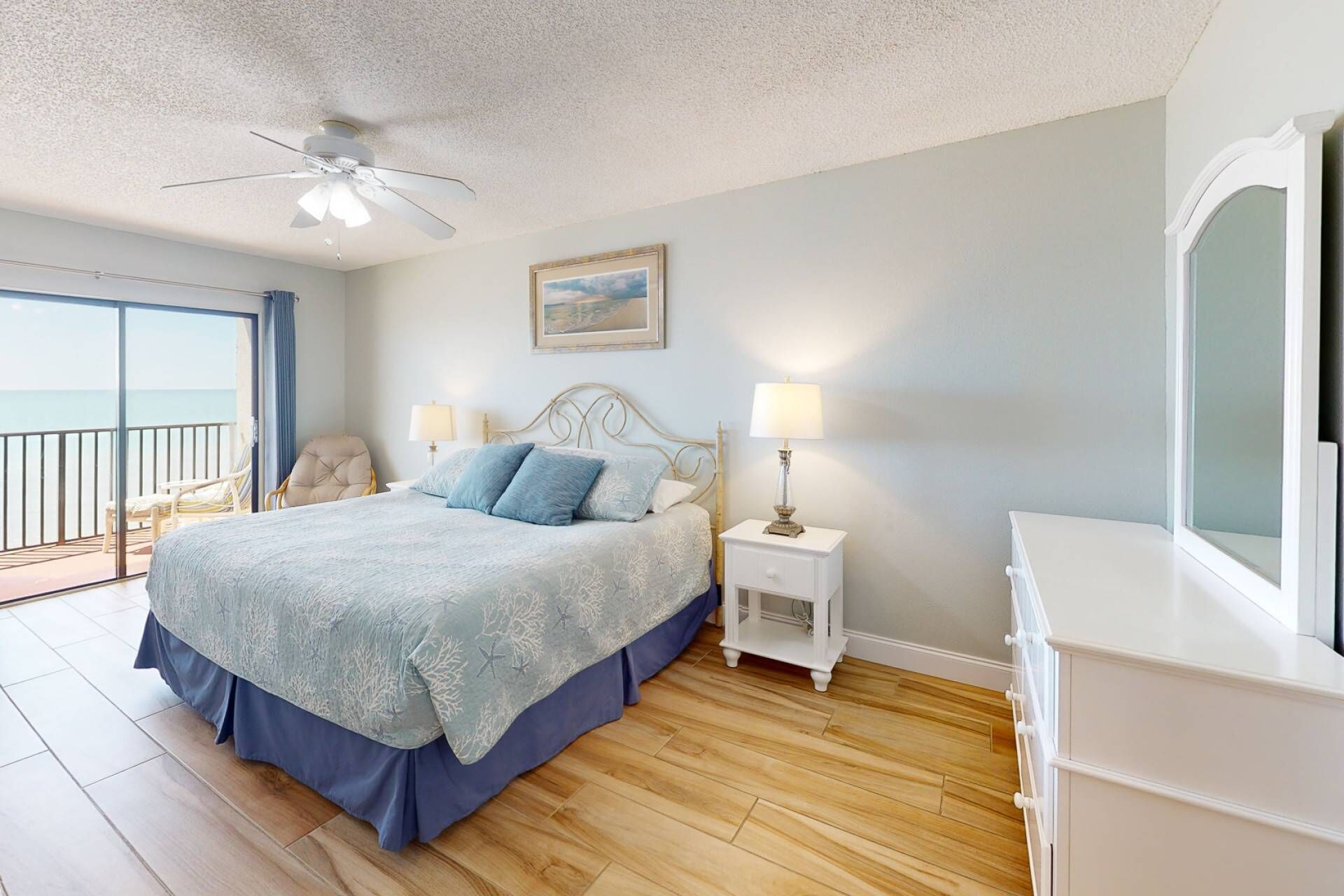 A bedroom with a bed, two bedside lamps, a ceiling fan, a framed picture, a chair, and a view of the ocean.