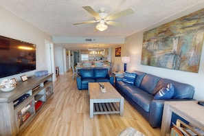 A spacious living room with a large flat-screen TV, a ceiling fan, and a dining area with a kitchen in the background.