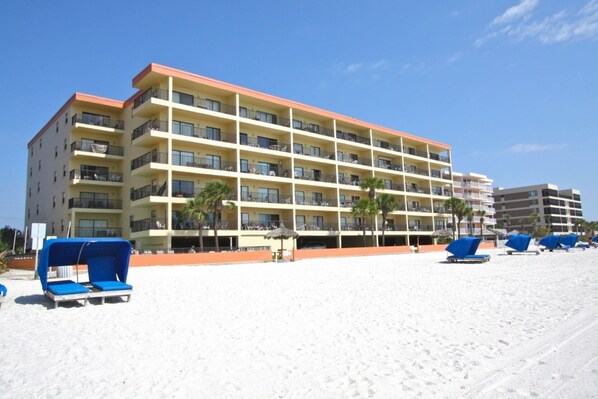 Ein mehrstöckiges Hotel mit Balkonen, einem Sandstrand davor und blauen Liegestühlen.