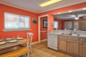 Une cuisine avec un coin repas, comprenant une fenêtre avec stores, une chaise blanche, une table à manger dressée avec des assiettes et des verres, et un îlot de cuisine doté d’un évier et de placards.