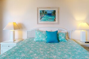 A bed with a blue and white starfish patterned bedspread, two matching pillows, and a framed tropical beach painting above it.