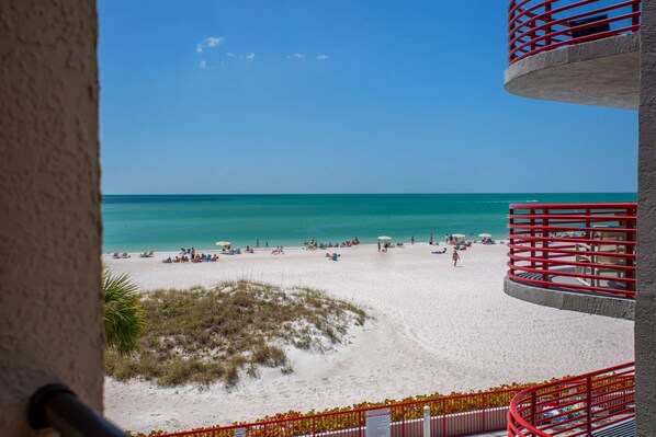 A beach view from a balcony with red railings, sandy beach, and clear blue water.