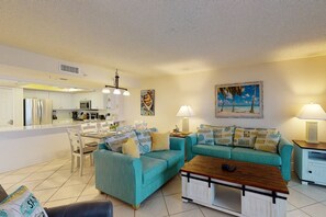 A modern living room with teal sofas, a wooden coffee table, and a kitchen area with white cabinets and a refrigerator.
