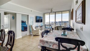 A spacious living area with a dining table, chairs, and a view of the city through large windows. - Beachside Biloxi Club Condo: Balcony w/ Ocean View (Biloxi)