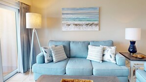 A living room with a light blue sofa, a floor lamp, a table with a lamp, and a beach-themed painting on the wall. - Unwind Together with a Beachfront View, Beach, Pool. 6th Floor. Tickets to Top Attractions Daily (Madeira Beach)