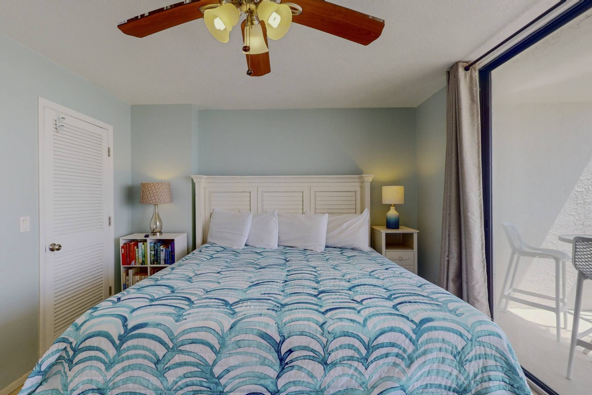 Un dormitorio con cama, ventilador de techo, mesita de noche, lámpara, estantería y ventana con cortinas.