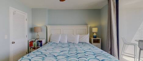 Un dormitorio con cama, ventilador de techo, mesita de noche, lámpara, estantería y ventana con cortinas.