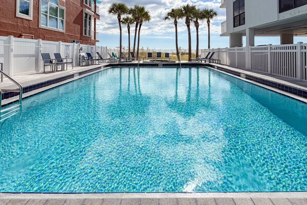 A rectangular swimming pool with clear blue water, surrounded by a white fence, and palm trees in the background.