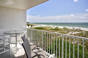 A balcony with a view of a beach, white railing, and two tables with chairs.