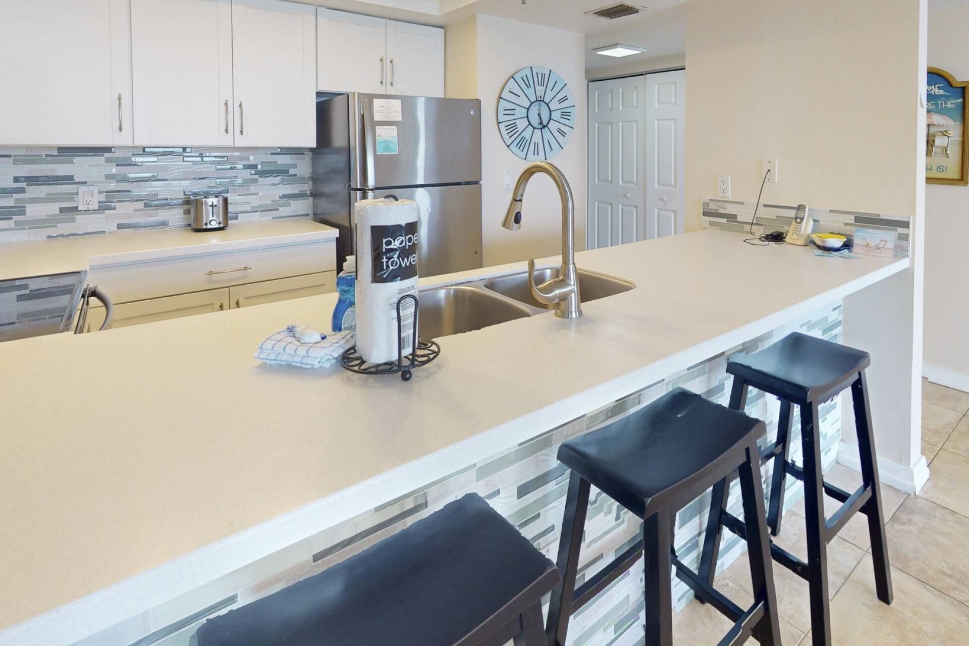 Een moderne keuken met een roestvrijstalen koelkast, een koffieapparaat en een spoelbak met een moderne kraan. Er staan twee barkrukken bij het eiland.