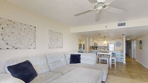 A spacious living room with a large white sofa, a ceiling fan, and a kitchen area in the background. - Incredible Beachfront View Private Balcony! 2 King Bedrooms, Pool & Hot Tub. Quiet and Relaxing Unit (Madeira Beach)