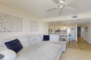 A spacious living room with a large white sofa, a ceiling fan, and a kitchen area in the background.
