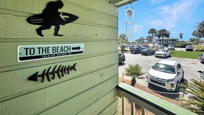 Un bâtiment sur lequel on peut lire l’inscription «DIRECTION LA PLAGE», une silhouette d’une personne tenant une planche de surf, et une vue sur des voitures garées et des palmiers.
