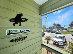 A building with a 'TO THE BEACH' sign, a silhouette of a person with a surfboard, and a view of parked cars and palm trees.