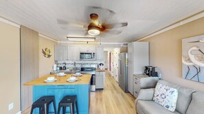 A modern kitchen with a central island, a dining area with stools, a refrigerator, and a living area with a sofa and wall art. - Great oceanfront 1st floor unit with beach, fishing pier, and pool access (Isle of Palms)