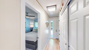 Un couloir mène à une chambre à coucher qui contient un lit, une table de chevet, une lampe et un ventilateur de plafond.