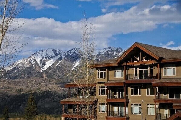 A multi-story building with balconies, set against a backdrop of snow-capped mountains and clear skies.