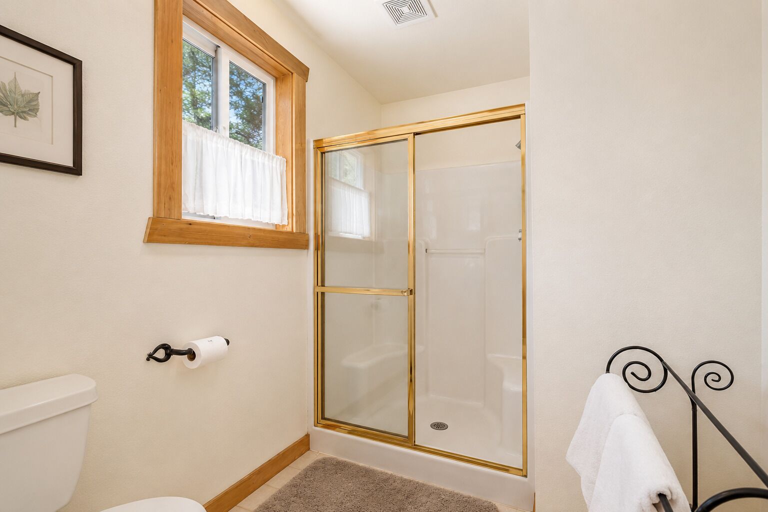 Une salle de bain comprenant des toilettes, une œuvre d’art représentant une feuille encadrée, une fenêtre avec des rideaux blancs, une douche avec un cadre doré et un porte-serviettes.