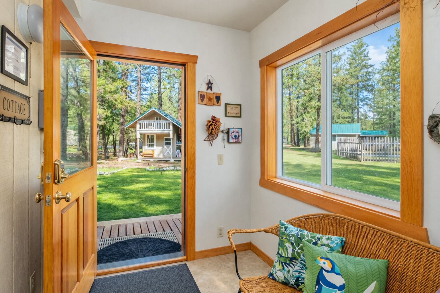A cozy entryway with a wooden door leading to a grassy yard, a wicker bench with colorful cushions, and a view of a house in the background.