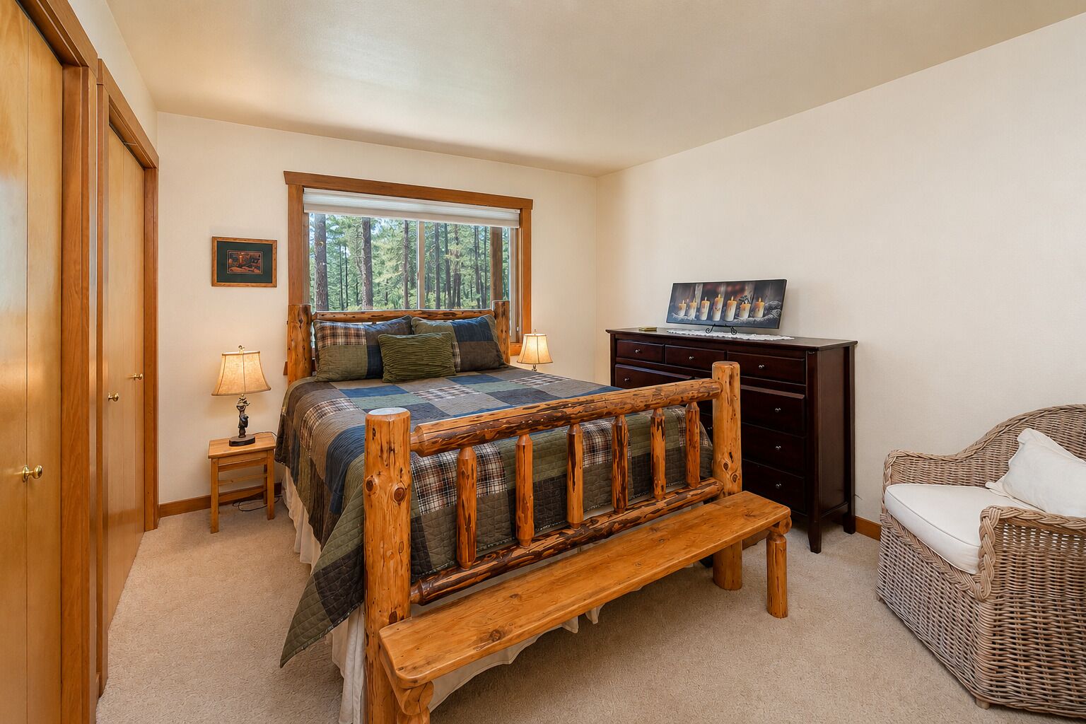 Un dormitorio con una cama de madera, una mesita de noche, un aparador, una silla de mimbre y un ventanal con vista al bosque.