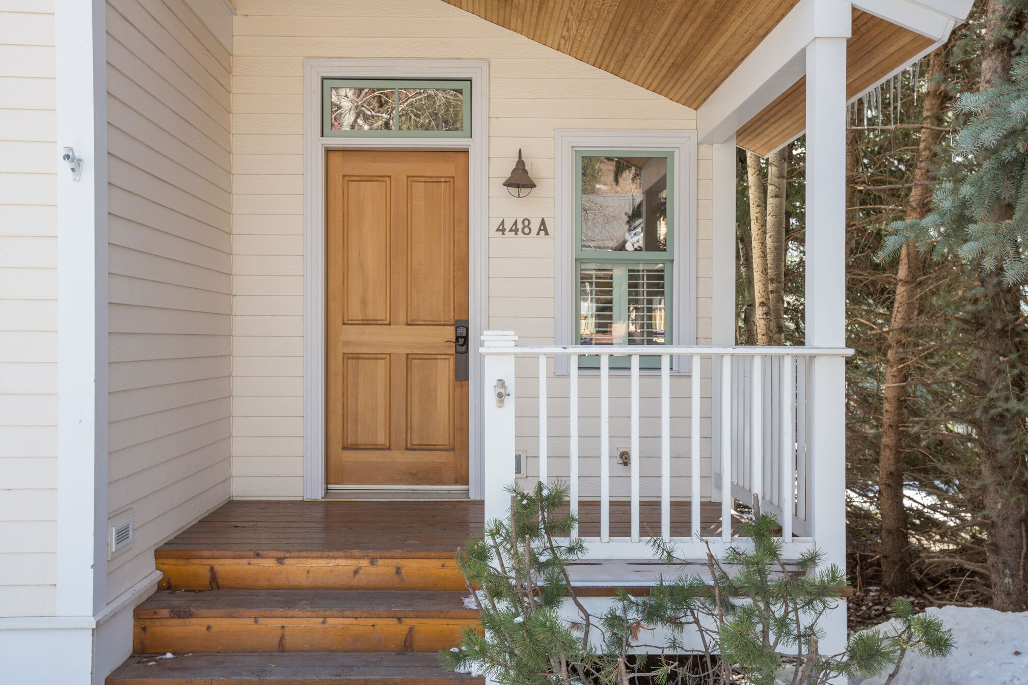 Une porte d’entrée en bois, avec le numéro « 448A » au-dessus, un porche blanc doté d’une balustrade et un petit arbre au premier plan.
