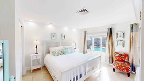 Un dormitorio con una cama, dos mesitas de noche, una lámpara auxiliar y una ventana que da a una piscina, además de una silla con un cojín de colores.
