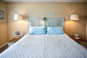 Quarto com cama, duas luminárias de cabeceira, um letreiro sobre a cama e uma mesinha com abajur.
