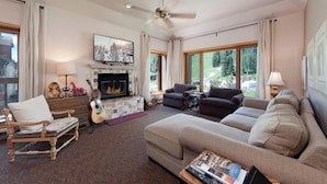 A living room with a fireplace, a ceiling fan, and a large window with a view of a green landscape.
