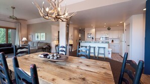 A spacious living area with a wooden dining table, blue chairs, a chandelier, and a kitchen in the background.