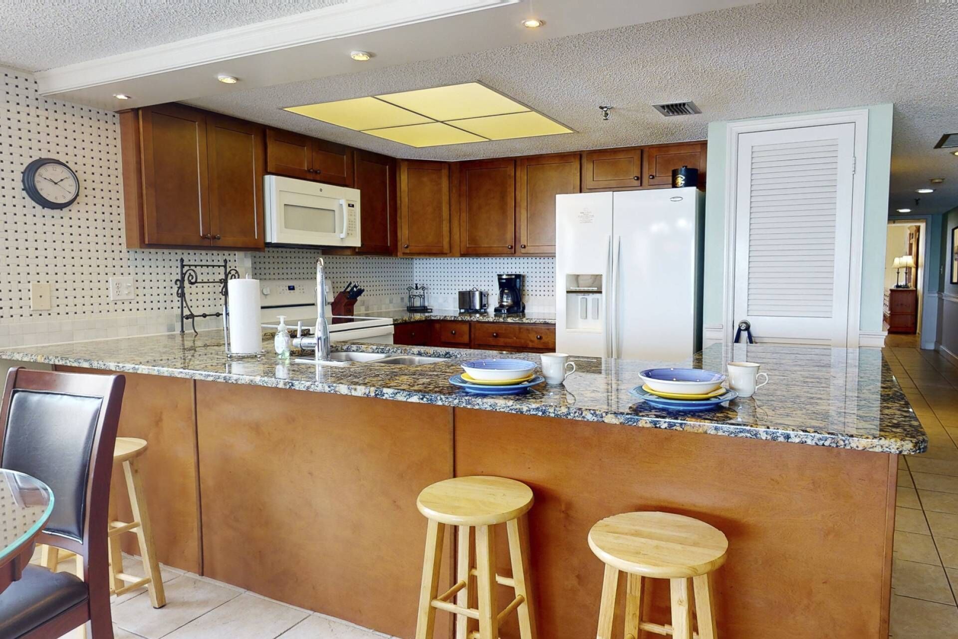 A modern kitchen with wooden cabinets, a white refrigerator, a microwave, and a granite countertop. There are two wooden stools and a glass table.