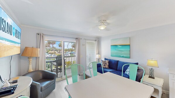 A room with a balcony, a sofa, a table, and a TV displaying a beach scene.