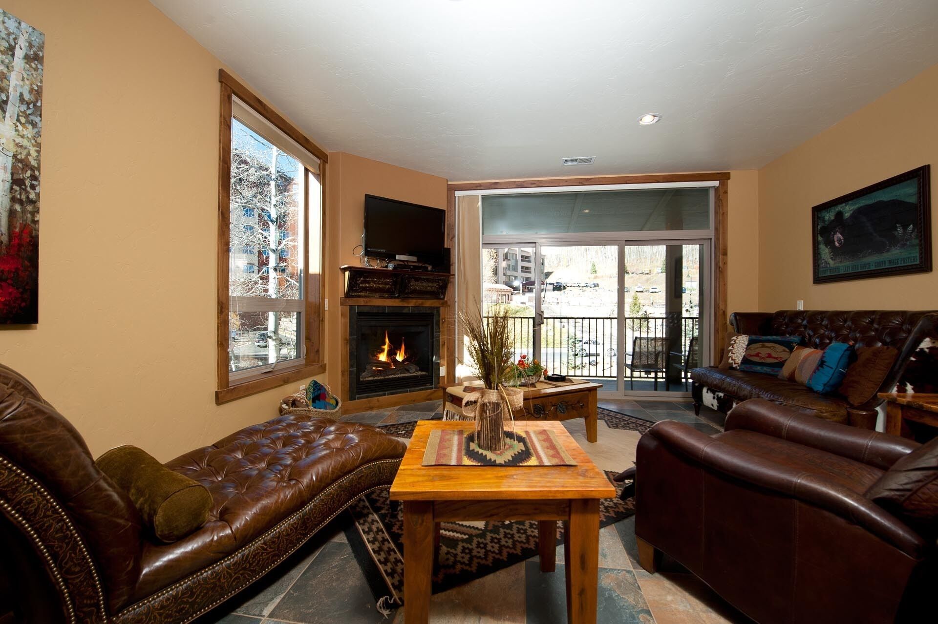 A cozy living room with a fireplace, leather sofas, a wooden coffee table, and a large window with a view of buildings.