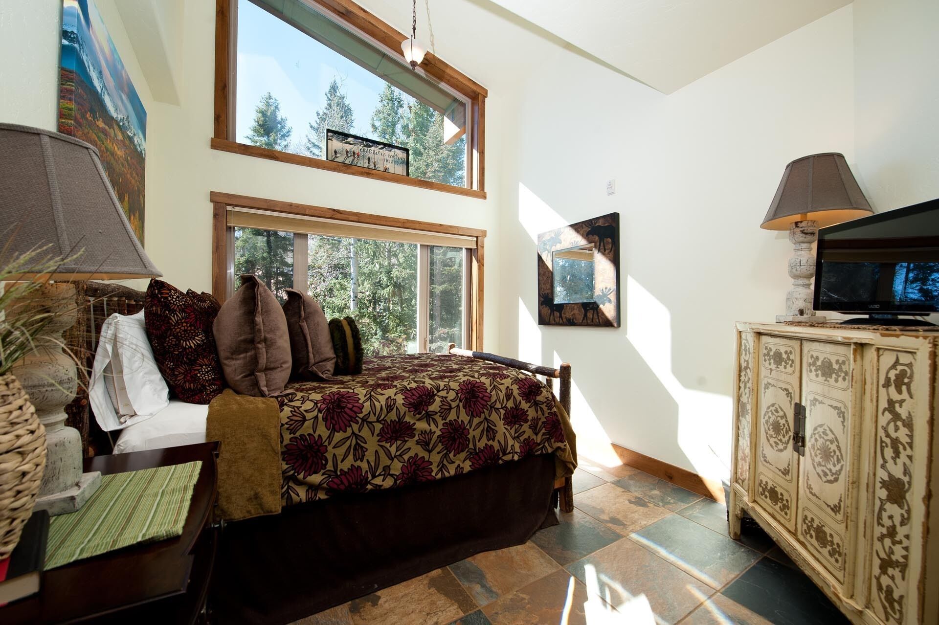 A bedroom with a large bed, a television, and a cabinet. There are windows providing natural light and a view of trees.