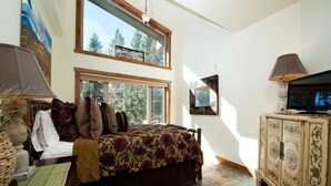 A bedroom with a large bed, a television, and a cabinet. There are windows providing natural light and a view of trees. - Professionally Decorated Peregrine Point Steps to Lifts (Durango)