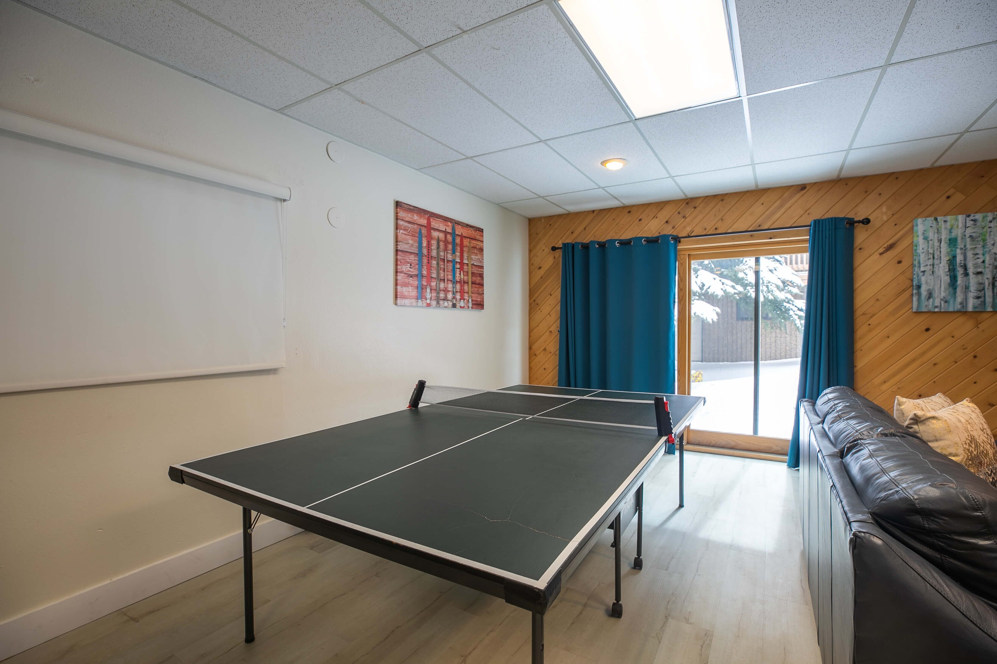 A ping pong table in a room with a wooden wall, a black leather couch, and a window with blue curtains.